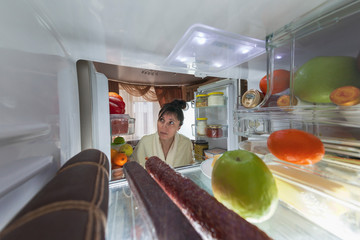 Beautiful brunette in a bathrobe is about to eat food near the fridge at night, toned