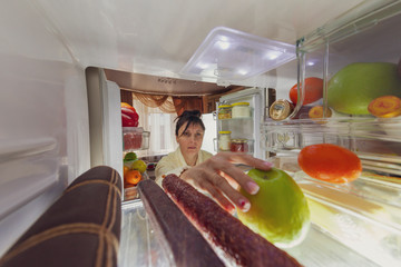 Beautiful brunette in a bathrobe is about to eat food near the fridge at night, toned