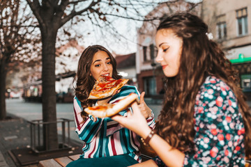 Two cheerful girl  talking and eating pizza outdoors, having fun together
