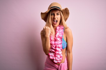 Young beautiful blonde woman on vacation wearing bikini and hat with hawaiian lei flowers angry and mad raising fist frustrated and furious while shouting with anger. Rage and aggressive concept.