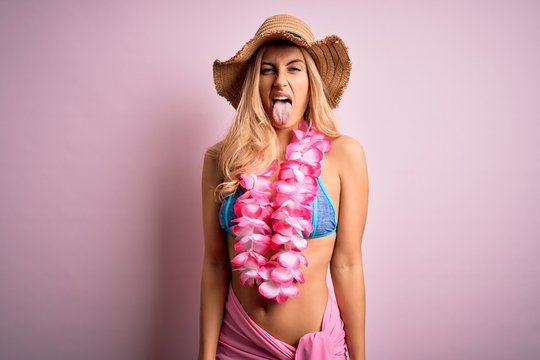 Young beautiful blonde woman on vacation wearing bikini and hat with hawaiian lei flowers sticking tongue out happy with funny expression. Emotion concept.