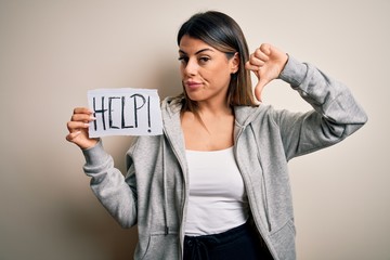 Young beautiful brunette woman holding paper with help message over white background with angry...
