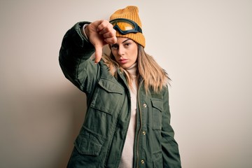 Young brunette skier woman wearing snow clothes and ski goggles over white background looking unhappy and angry showing rejection and negative with thumbs down gesture. Bad expression.