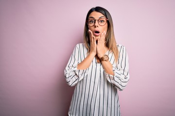 Young beautiful woman wearing casual striped t-shirt and glasses over pink background afraid and shocked, surprise and amazed expression with hands on face
