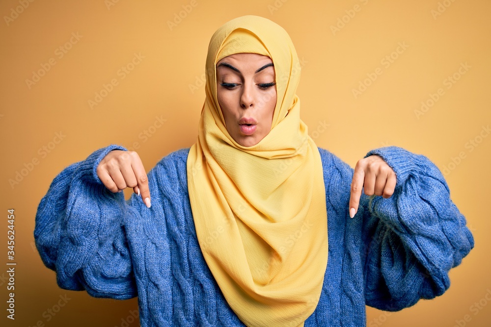 Canvas Prints Young beautiful brunette muslim woman wearing arab hijab over isolated yellow background Pointing down with fingers showing advertisement, surprised face and open mouth