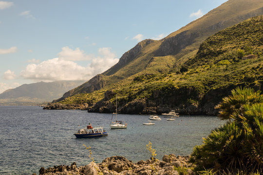 Mediterranean landscape of Sicily, Italy. View shot in Zingaro Nature Reserve. 
In the background boats, yachts. Mix of azur water, rich people, rock, coast, mountain, plants, sea, flora and sailing. 