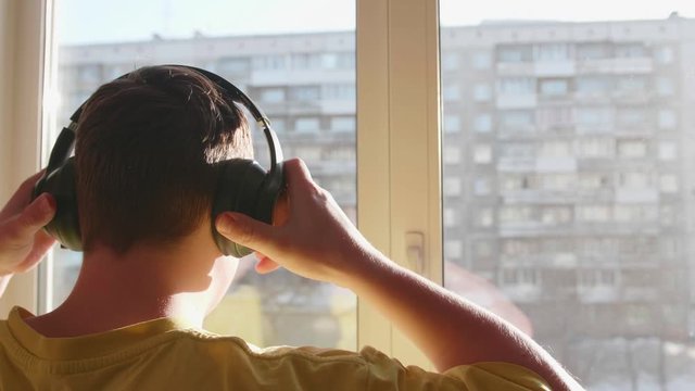 A young man stands near the window and puts on headphones to listen to music. Blurred background with sunset, teen enjoying music in headphones.