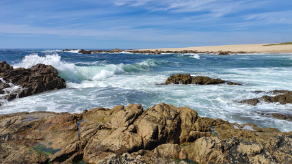 Fototapeta na wymiar Waves breaking over rocks at beach on bright sunny summer day in Povoa de Varzim, Portugal