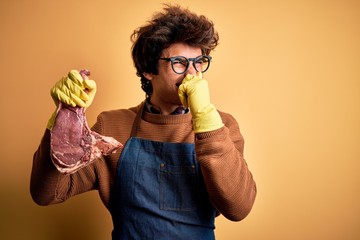 Young handsome butcher man holding meet steak standing over isolated yellow background smelling...