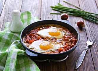 Shakshuka or fried eggs in tomato sauce with red bell pepper in a serving cast-iron frying pan on a wooden background. Village breakfasts. Israeli food.
