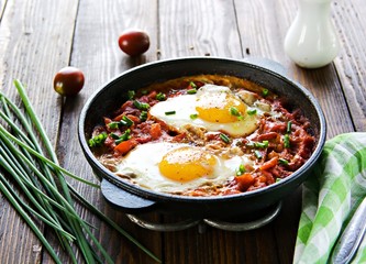 Shakshuka or fried eggs in tomato sauce with red bell pepper in a serving cast-iron frying pan on a wooden background. Village breakfasts. Israeli food.