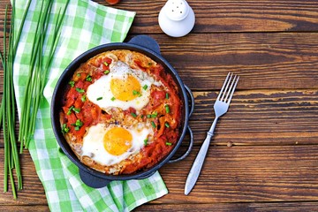 Shakshuka or fried eggs in tomato sauce with red bell pepper in a serving cast-iron frying pan on a wooden background. Village breakfasts. Israeli food.