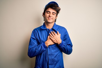 Young mechanic man wearing blue cap and uniform standing over isolated white background smiling with hands on chest with closed eyes and grateful gesture on face. Health concept.