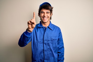 Young mechanic man wearing blue cap and uniform standing over isolated white background showing and pointing up with finger number one while smiling confident and happy.