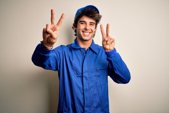 Young mechanic man wearing blue cap and uniform standing over isolated white background smiling looking to the camera showing fingers doing victory sign. Number two.