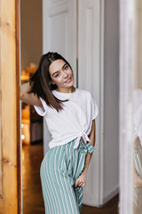Fototapeta na wymiar Enchanting female model in trendy pants enjoying photoshoot at home. Indoor portrait of inspired brunette woman in casual t-shirt relaxing in her apartment.