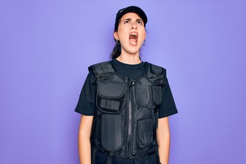 Young police woman wearing security bulletproof vest uniform over purple background angry and mad screaming frustrated and furious, shouting with anger. Rage and aggressive concept.