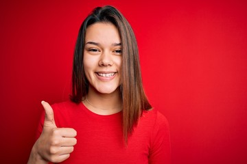 Young beautiful brunette girl wearing casual t-shirt over isolated red background doing happy thumbs up gesture with hand. Approving expression looking at the camera showing success.