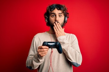 Young gamer man with curly hair and beard playing video game using joystick and headphones cover mouth with hand shocked with shame for mistake, expression of fear, scared in silence, secret concept