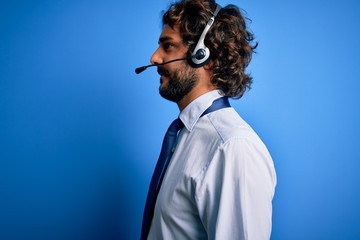 Young handsome call center agent man with beard working using headset over blue background looking...