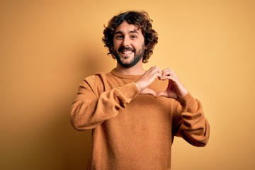 Fototapeta premium Young handsome man with beard wearing casual sweater standing over yellow background smiling in love doing heart symbol shape with hands. Romantic concept.