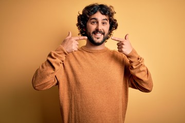 Young handsome man with beard wearing casual sweater standing over yellow background smiling cheerful showing and pointing with fingers teeth and mouth. Dental health concept.