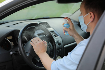 Spraying antibacterial sanitizer spray on steering wheel car, infection control concept. Prevent Coronavirus, COVID-19, flu. Man wearing in medical protective mask driving a car. Disinfecting wipes.
