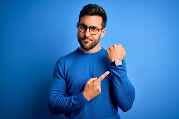 Young handsome man with beard wearing casual sweater and glasses over blue background In hurry pointing to watch time, impatience, looking at the camera with relaxed expression - obrazy, fototapety, plakaty