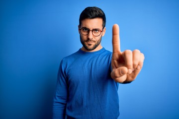Young handsome man with beard wearing casual sweater and glasses over blue background Pointing with finger up and angry expression, showing no gesture - obrazy, fototapety, plakaty