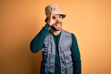 Handsome tourist man with beard on vacation wearing explorer hat over yellow background doing ok gesture with hand smiling, eye looking through fingers with happy face.
