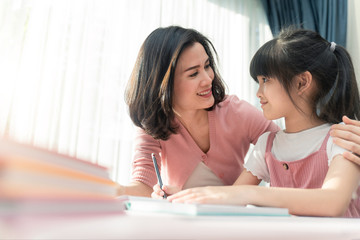 Fototapeta premium Homeschool Asian young little girl learning, reading and does homework with kind mother help, teach and encourage. Mom hug daughter and smile. Girl happy to study at home education together with mom.