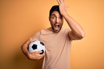 Handsome player man with beard playing soccer holding footballl ball over yellow background...