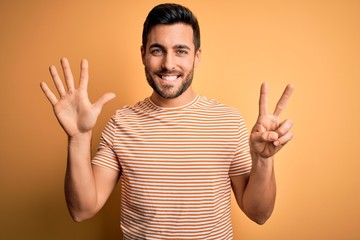 Young handsome man with beard wearing casual striped t-shirt over yellow background showing and pointing up with fingers number seven while smiling confident and happy.