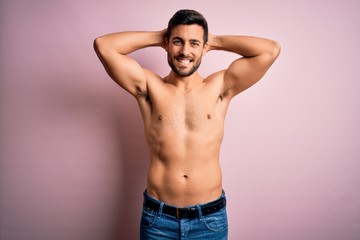 Young handsome strong man with beard shirtless standing over isolated pink background relaxing and stretching, arms and hands behind head and neck smiling happy