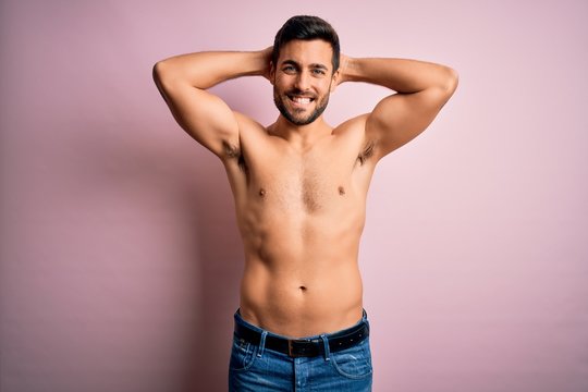 Young handsome strong man with beard shirtless standing over isolated pink background relaxing and stretching, arms and hands behind head and neck smiling happy