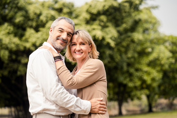 Fototapeta premium Senior elder Caucasian couple sitting on ground together in park in Autumn. Wife hugging husband from behind and smile with happiness. Beautiful love relation and care of retirement old people.