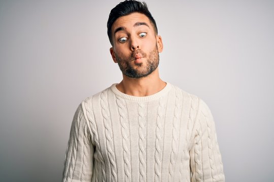 Young handsome man wearing casual sweater standing over isolated white background making fish face with lips, crazy and comical gesture. Funny expression.