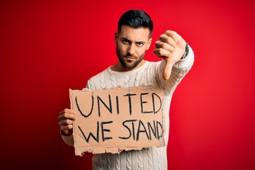 Young handsome activist man protesting holding cardboard with unity message with angry face,...