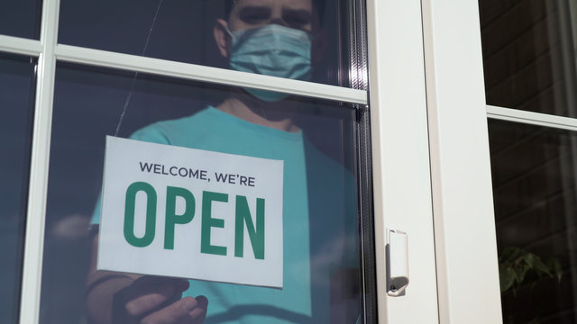 Close Up Of The Male Caucasian Hands Turning A Signboard On The Glass Door Of The Shop From CLOSED To OPEN. Reopening Of A Small Business Activity After Quarantine Covid-19 Emergency.