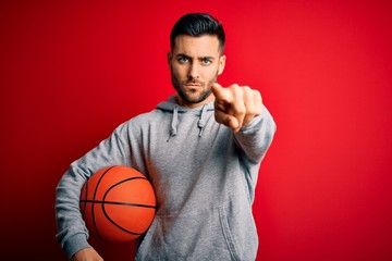 Young sports man holding basketball ball over red isolated background pointing with finger to the camera and to you, hand sign, positive and confident gesture from the front