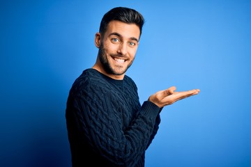 Young handsome man wearing casual sweater standing over isolated blue background pointing aside with hands open palms showing copy space, presenting advertisement smiling excited happy