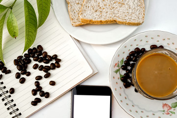 Close up of coffee beans on notebook,two pieces of bread,mobile phone,cup of coffee with saucer.