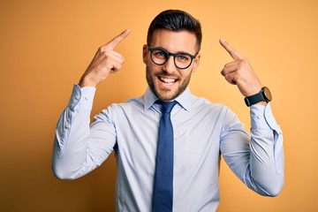 Young handsome businessman wearing tie and glasses standing over yellow background smiling pointing...