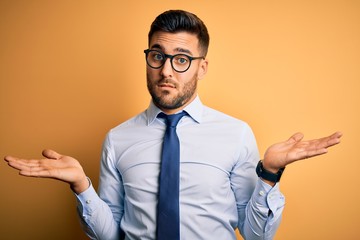 Young handsome businessman wearing tie and glasses standing over yellow background clueless and confused with open arms, no idea concept.