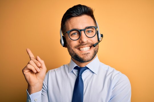 Young business operator man wearing customer service headset from call center with a big smile on face, pointing with hand and finger to the side looking at the camera.