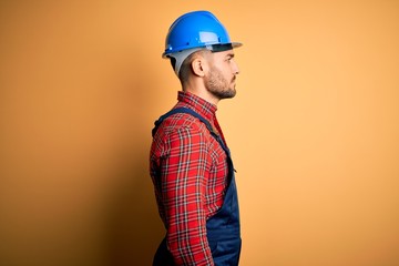 Young builder man wearing construction uniform and safety helmet over yellow background looking to side, relax profile pose with natural face with confident smile.