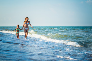 Young pretty mother and little daughter run on the sea shore during vacation on a sunny warm summer day against a blue sky. Concept vacation with children and wellness
