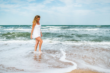 Charming young woman in a white dress walks along the calm sea waves on the sandy coast against a background of blue sky. White clouds on a sunny warm summer day