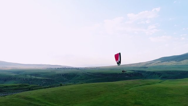 A person with a red, black and white parachute flying in a blue sky above green fields surrounded by a highway and electric wires 