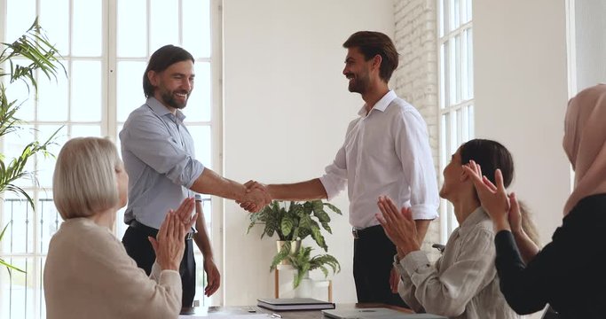 Happy diverse multiracial older and younger colleagues watching male boss supervisor shaking hands, praising intern coworker in office. Smiling employees clapping hands, supporting successful worker.